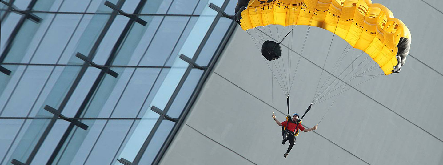 A skydiver wearing a red shirt maneuvers his yellow parachute near a modern building.