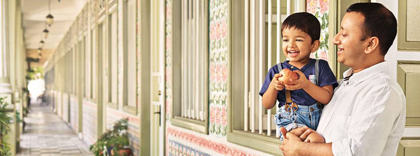 A man standing in a long outdoor corridor, holds a young boy who is gripping an apple in his hands.