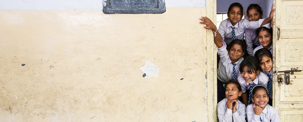 Smiling and laughing, uniformed Indian schoolchildren look out the door of a rural school. 